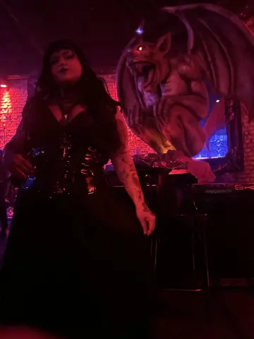 Woman in gothic black corset dress posing next to a large gargoyle statue in moody red lighting.