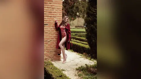 Elegant woman in a red coat and lace bodysuit posing by a rustic brick wall in a lush green garden.