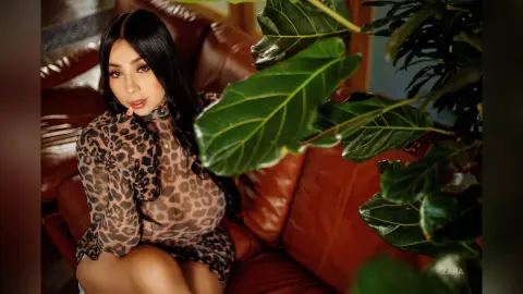 Young woman in a leopard print blouse sitting on a brown leather sofa near lush green indoor plants.