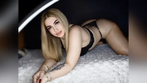 ZaraPlaya poses on a white textured bed, wearing black lace lingerie and showing her long blonde hair and arm tattoo.