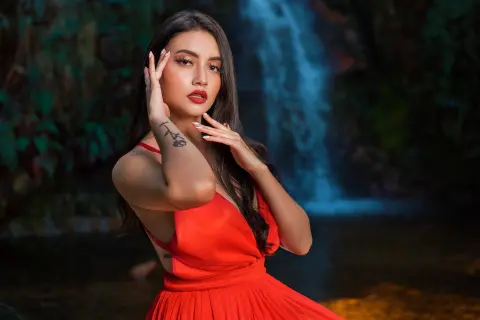 Elegant woman in a red dress posing by a tropical waterfall in a lush, moody jungle setting.