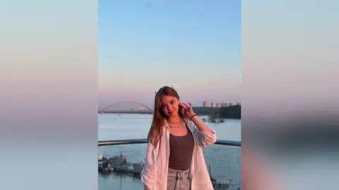PaulettaRerucha stands on a balcony overlooking a river, wearing a grey tank top and an open white shirt.