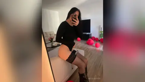 Woman in a black long-sleeve bodysuit and lace stockings posing in a modern living room with pink balloons.