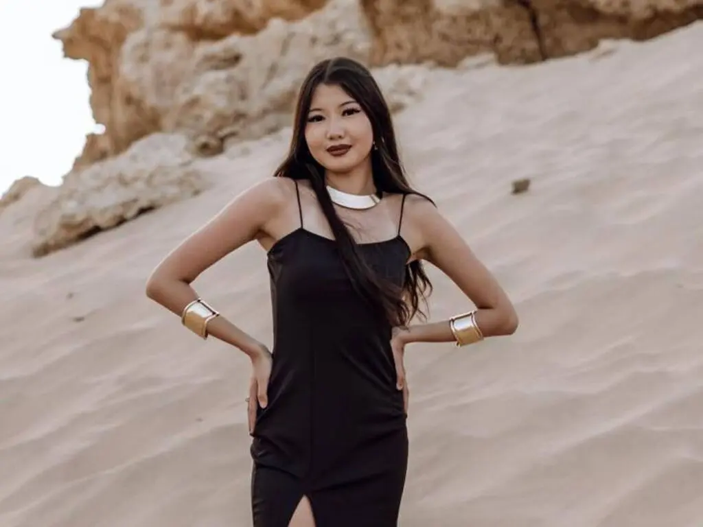 Smiling young woman in a black slip dress and gold cuffs posing on a sunny sand dune landscape.