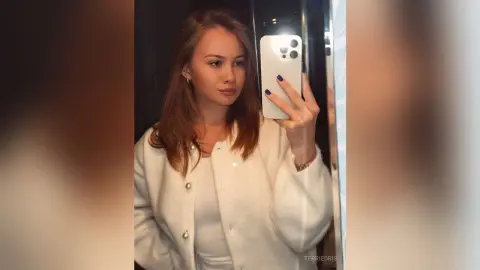 Young woman in a white fuzzy cardigan taking a stylish mirror selfie in a modern setting.