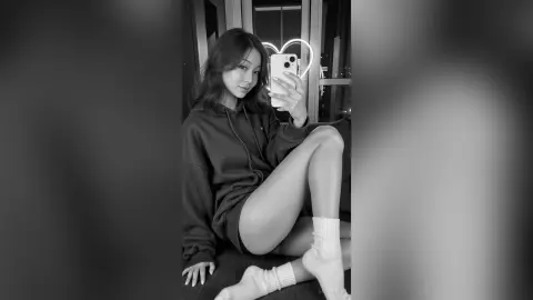 LeahReel — Young woman in a cozy dark hoodie sitting by a window with a heart light in a moody black and white setting.