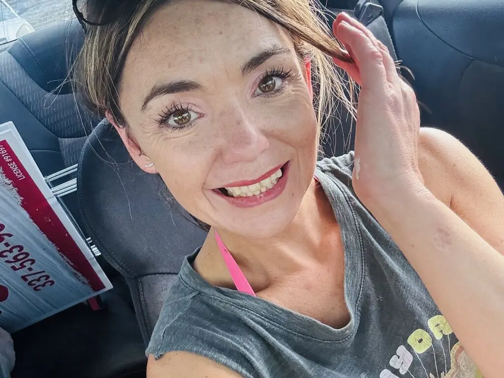 Smiling woman in a grey tank top relaxing in a car on a sunny day.