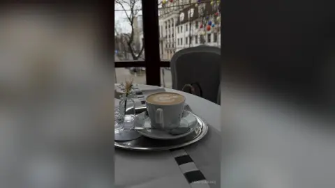 A warm cup of latte coffee on a cafe table with a view of city buildings through a window.