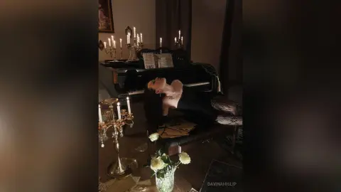 Moody portrait of a woman in a black dress reclining near a grand piano and lit candles in a dark room.