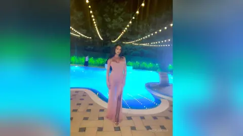 NicoleBella stands poolside at night wearing a long, tight pink dress with a low-cut neckline.