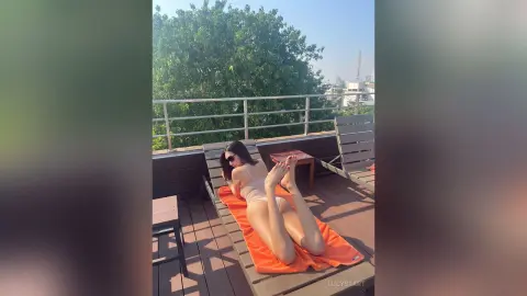 Relaxed woman in a beige bikini sunbathing on an orange lounge chair on a sunny rooftop balcony.