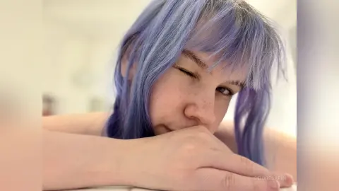 Young woman with lavender purple hair winking playfully in a bright, cozy indoor setting.