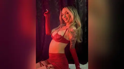 Smiling blonde woman in red lingerie and long gloves posing in moody, dim lighting.