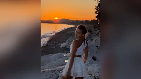 Young woman in a white summer outfit standing on a rocky beach during a golden sunset.