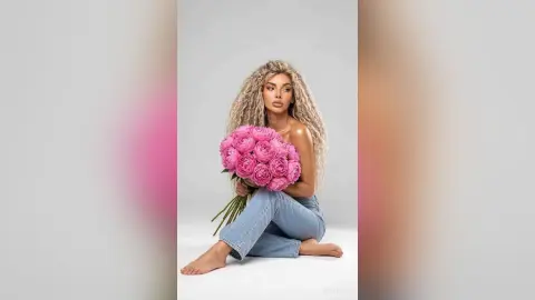 Blonde woman in denim jeans sitting with a large bouquet of pink roses against a white studio background.