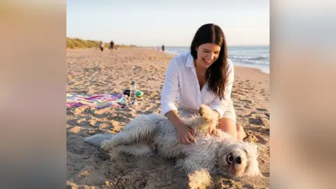 Smiling woman in a white linen shirt petting a fluffy white dog on a sunny sandy beach.