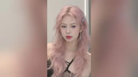 Young woman with wavy pink hair wearing a black mesh top in a soft, dreamy indoor setting.