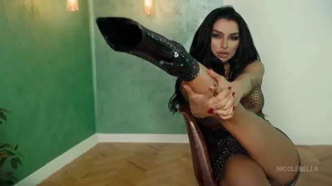 NicoleBella sits on a chair, lifting one leg up to show her bare thigh and foot, wearing a black mesh bodysuit.