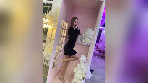 Elegant woman in a chic black dress posing in a dreamy pink floral photo booth setting.