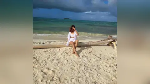 Woman in white summer outfit sitting on a driftwood log at a tropical beach under a cloudy sky.