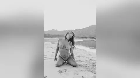 Black and white portrait of a woman in a bikini relaxing on a sandy beach under a bright sky.