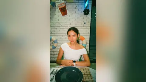 Young woman in a white t-shirt sitting at a cafe table with a relaxed mood against a white brick wall.