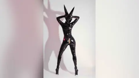 Woman in a sleek black latex bodysuit and high heels posing against a minimalist pink and white studio background.