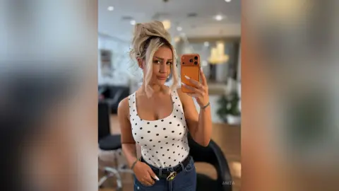 Anita poses for a mirror selfie wearing a white polka-dot tank top and blue denim jeans.