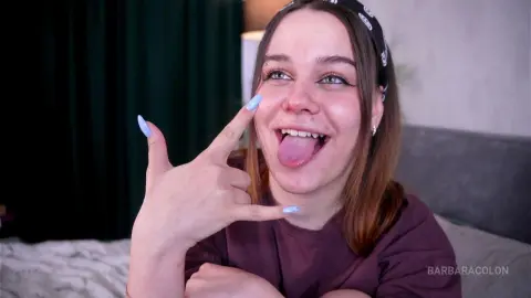Cheerful young woman with long brown hair and purple nails making a hand gesture in a cozy bedroom setting.