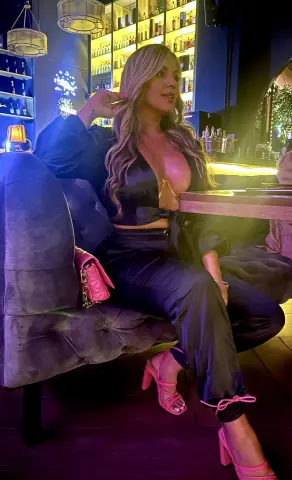 Glamorous woman in a black satin outfit sitting on a velvet sofa in a moody, dimly lit lounge bar.