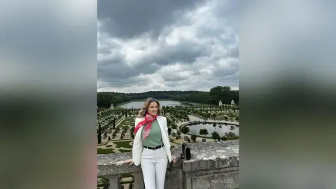 MayaDesi poses in a white blazer and green top against a cloudy sky overlooking a large garden and lake.
