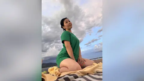 Smiling woman in a green summer dress sitting on a beach towel under a cloudy sky.