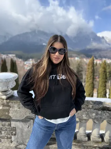 Young woman in a black hoodie and blue jeans posing outdoors against a scenic mountain landscape.