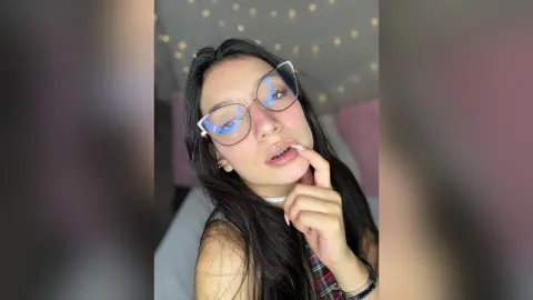 AleDilucas poses close-up wearing clear glasses and a white choker, touching her lip with her hand while looking at the camera.