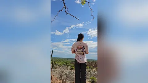 RossiNoleen stands outdoors with her back to the camera, wearing a white t-shirt and black pants against a blue sky.