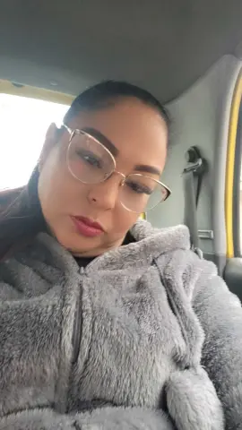 Woman with glasses wearing a cozy grey faux fur jacket sitting inside a car with a relaxed mood.