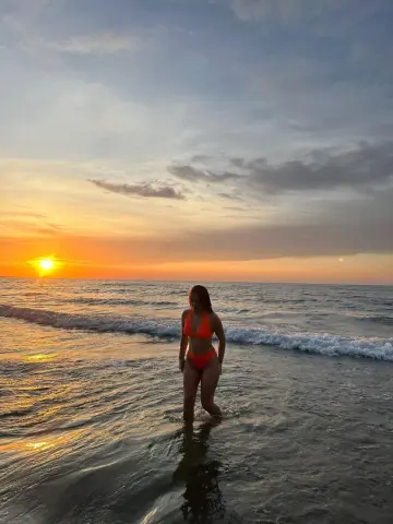 Woman in an orange bikini standing in ocean waves during a golden sunset at the beach.