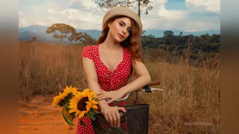 ValeryRouge poses in a red polka-dot sundress while holding bright sunflowers next to a bicycle in a grassy field.