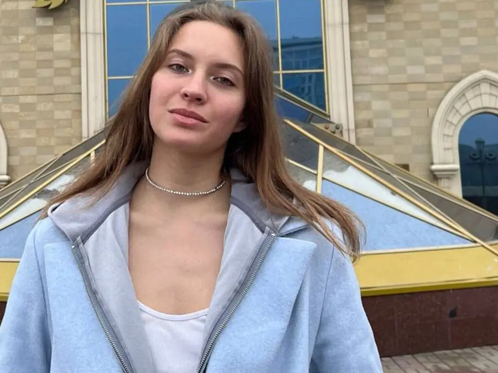 Smiling young woman in a light blue jacket and silver necklace standing before a modern glass building.