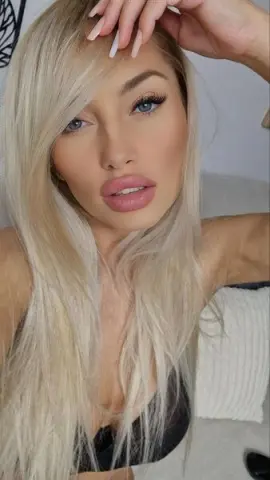 Blonde woman with glamorous makeup and long hair wearing a black lace top in a bright indoor setting.
