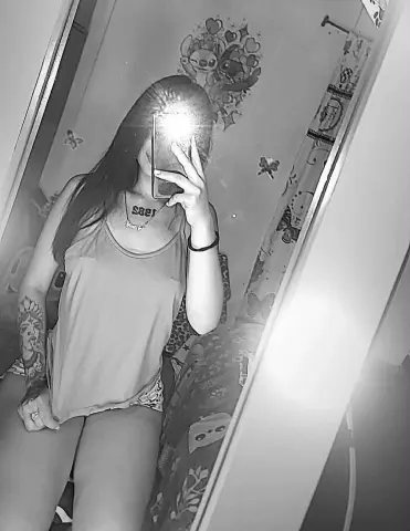 Black and white mirror selfie of a young woman in a tank top with long hair in a bedroom setting.