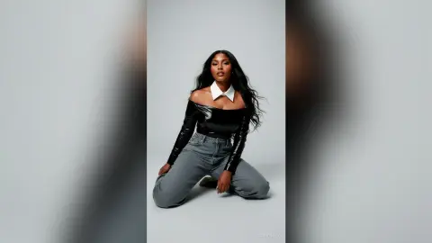 Stylish Black woman in a black off-the-shoulder top and grey jeans posing confidently against a grey backdrop.