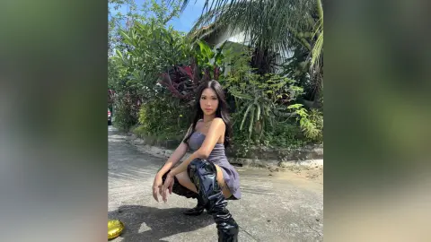Stylish woman in a grey dress and black patent leather boots posing in a lush tropical garden setting.