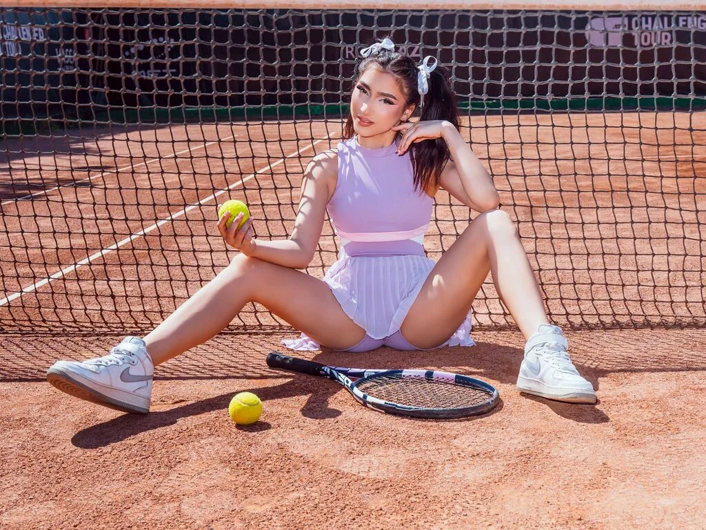 NickiHart sits on a clay tennis court in a white sleeveless bodysuit, holding a tennis ball near her leg.