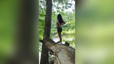 Young woman in a pink top and shorts standing on a fallen log in a lush green forest setting.