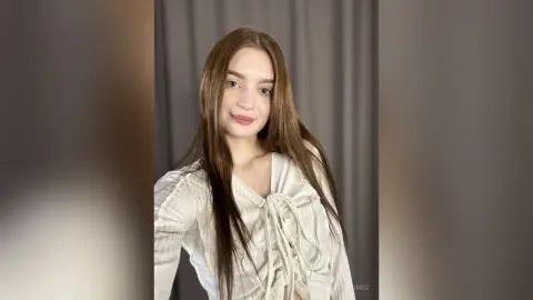 Young woman with long brown hair wearing a white long-sleeve blouse in a minimalist studio setting.