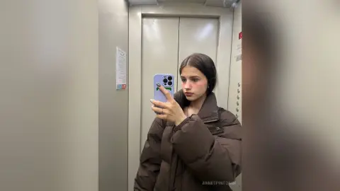 Young woman in a brown puffer jacket taking a mirror selfie in a modern elevator.
