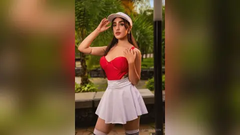 Young woman in a red crop top and white mini skirt posing outdoors in a lush green garden setting.