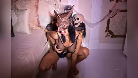 Woman in black mesh lingerie wearing a bronze demon mask crouching in a luxurious bedroom setting.