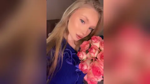 ViolaFausel poses with a large bouquet of pink roses, wearing a blue ribbed top with ruffles and showing her long blonde hair.